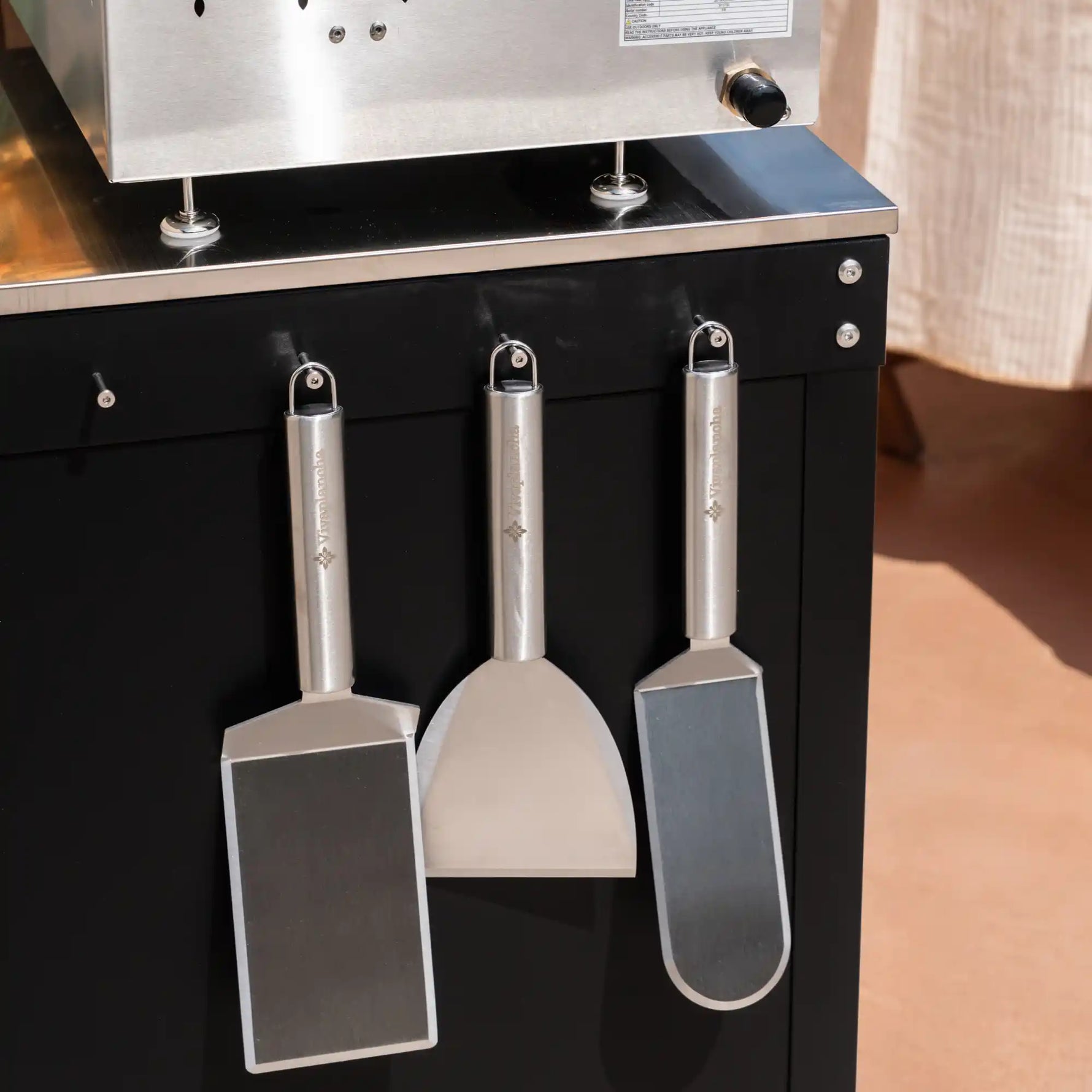  Gros plan sur trois ustensiles de cuisine (spatules Vivaplancha) suspendus sur le flanc de la desserte Bodega, sous la plancha.