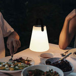 Lampe conique solaire allumée, diffusant une lumière chaude sur une table dressée pour un dîner en extérieur, entourée de plats et de convives.