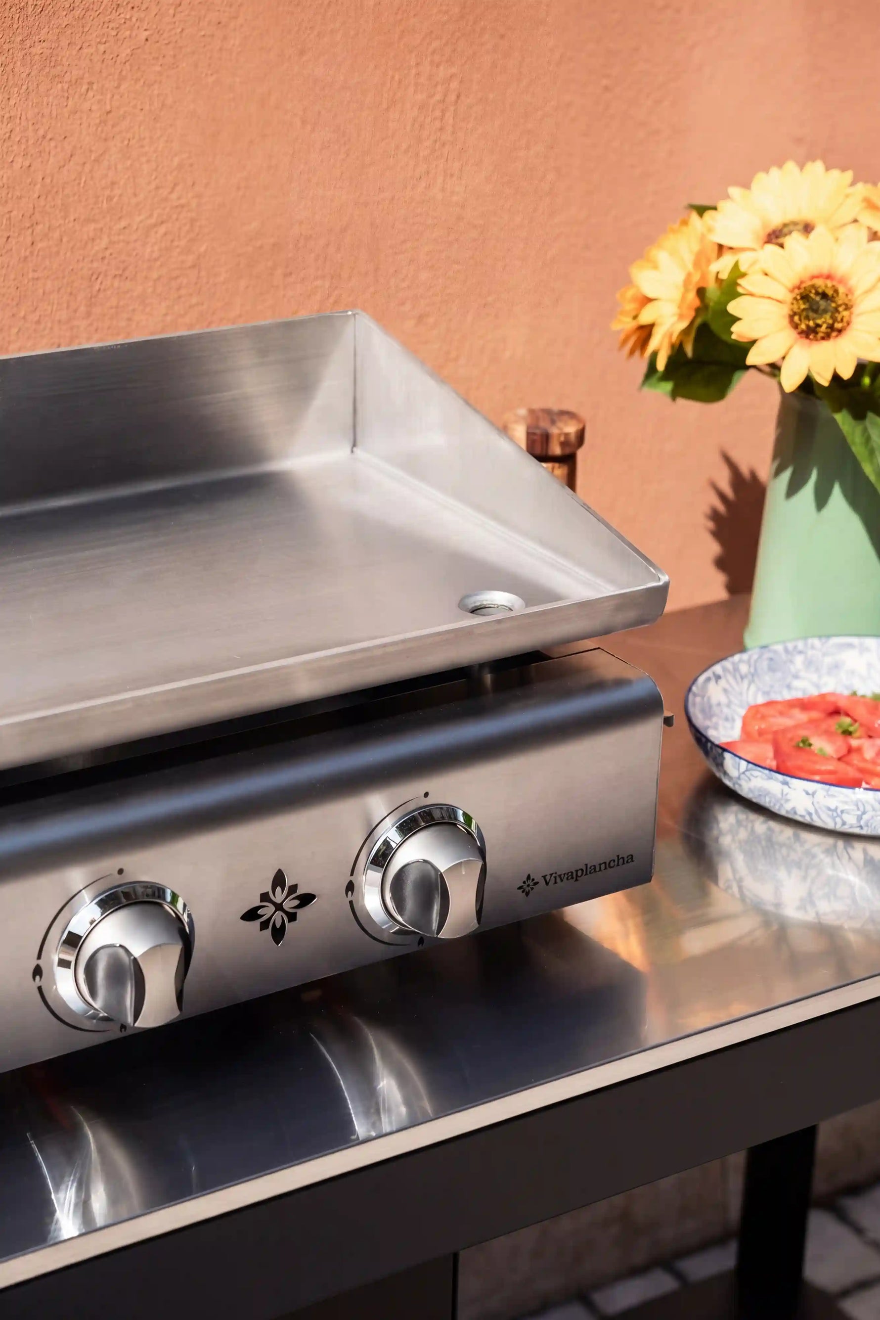 Plancha en inox, sans revêtement, posée sur un support extérieur, avec une touche de fleurs jaunes