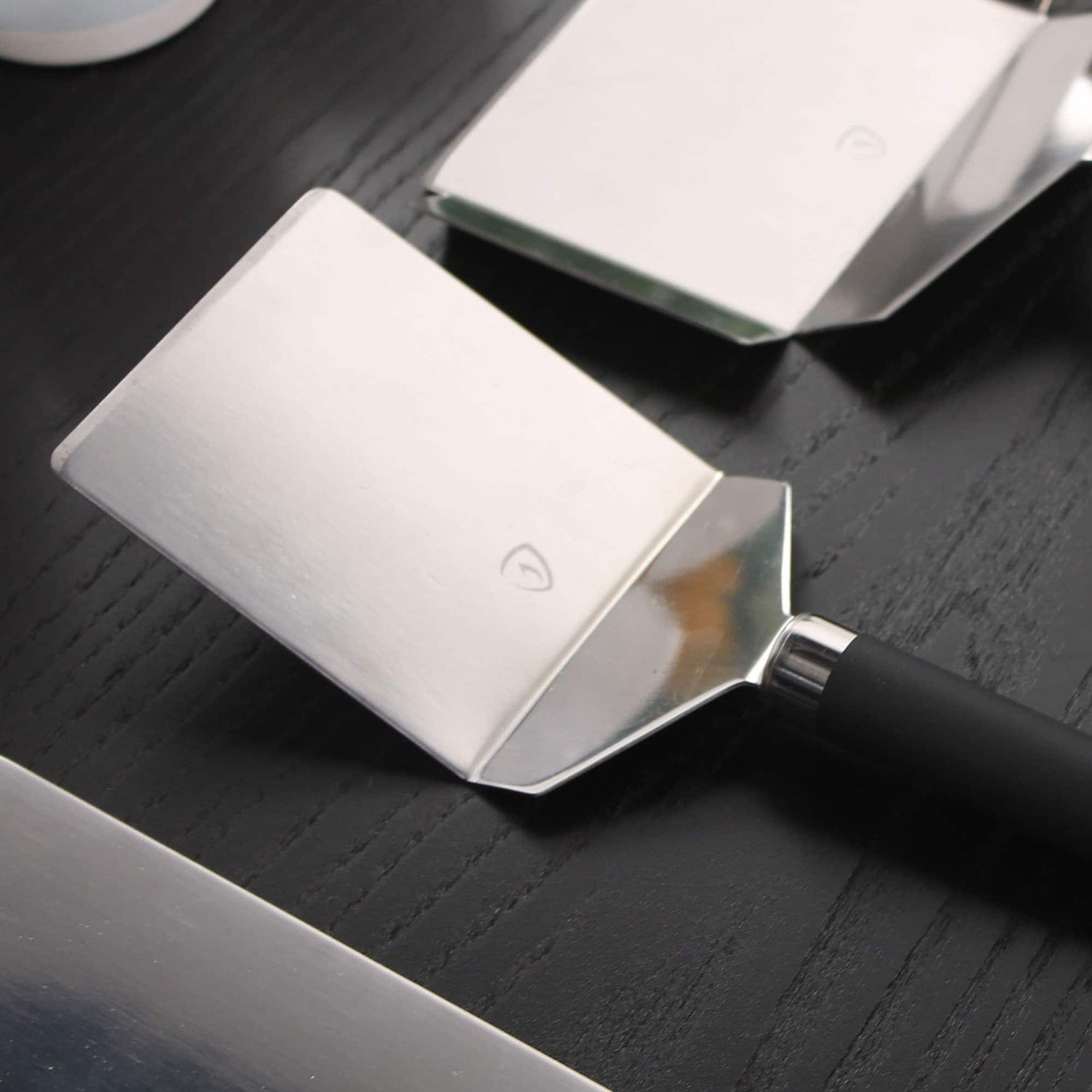 Spatules de cuisine en inox avec manche noir, posées sur un plan de travail sombre, montrées en gros plan.