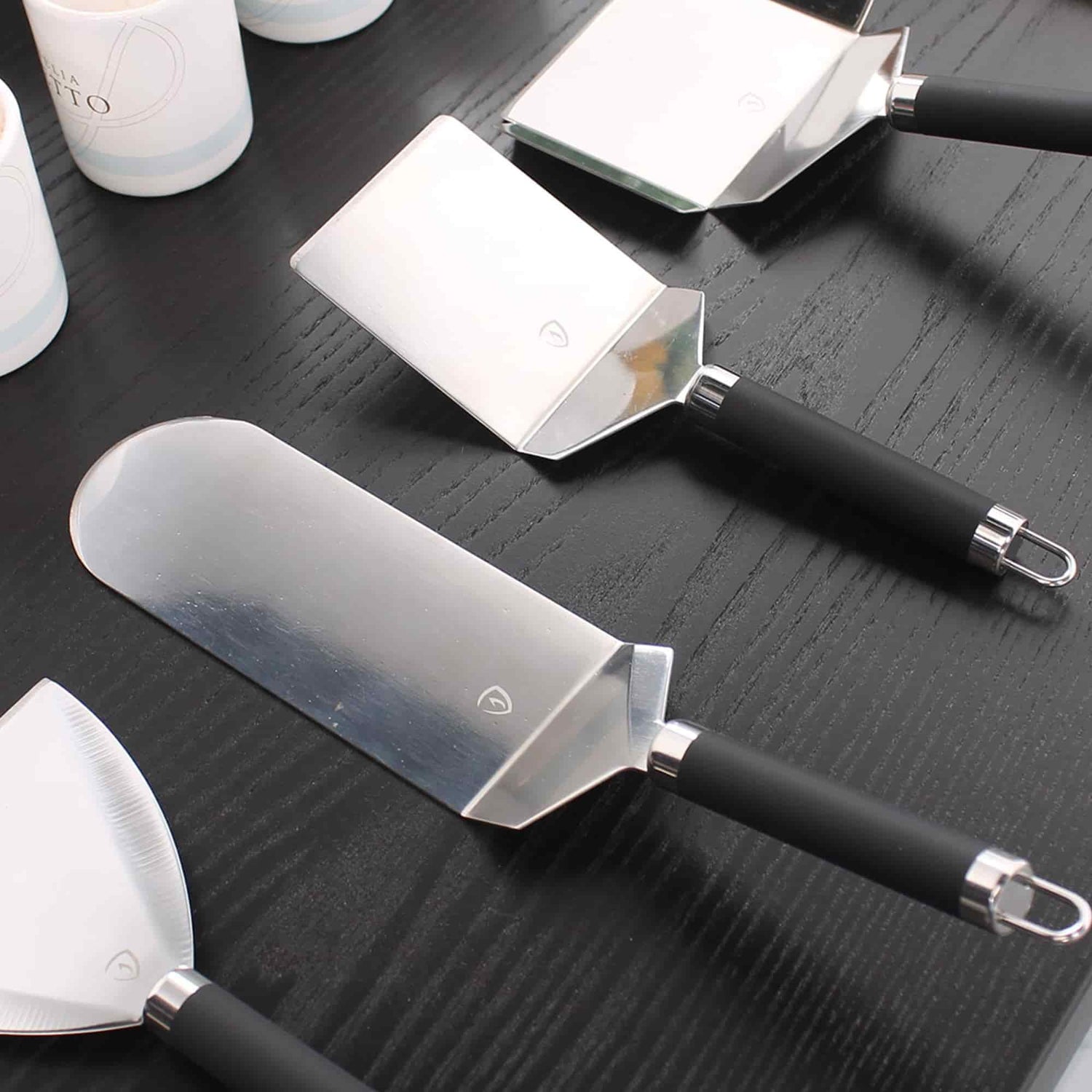 Ensemble de spatules en inox avec manches noirs, disposées sur un plan de travail en bois foncé, montrées en vue rapprochée.