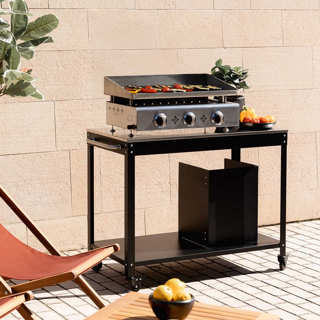 Plancha gaz Vivaplancha Pimientos 3 feux en inox, installée sur une desserte extérieure noire, en cuisson avec des légumes grillés.